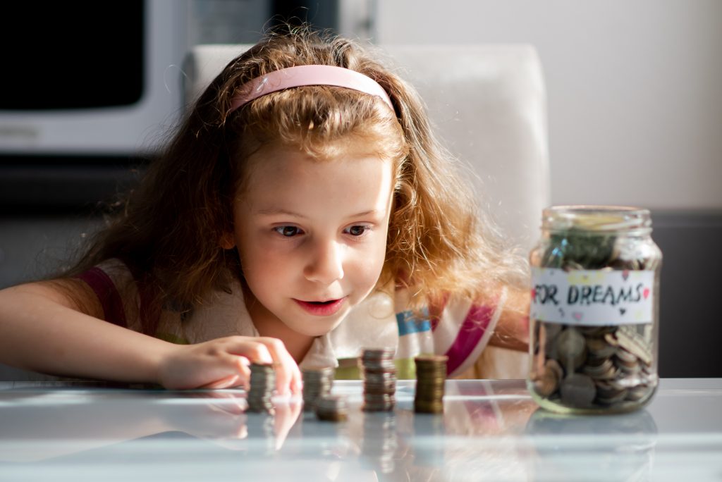 A sweet girl with full piggy bank of coins. On the piggy bank there is an inscription for dream. The girl is at home. She sits at a table and counts the coins. The money. Savings. Childhood.