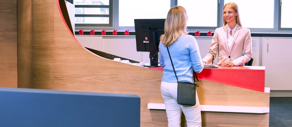 Blonde Kundenberaterin empäfngt Kundin am ServicePoint. Die Kundenberaterin lächelt freundlich.