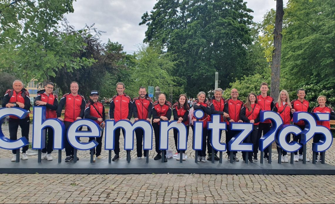 Eine Gruppe von Menschen in passenden rot-schwarzen Sportjacken steht hinter einem großen „Chemnitz 2025“-Schild in einer Parkanlage.
