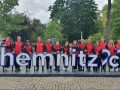 Eine Gruppe von Menschen in passenden rot-schwarzen Sportjacken steht hinter einem großen „Chemnitz 2025“-Schild in einer Parkanlage.