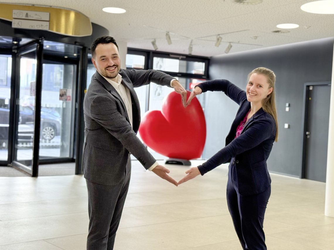 Carina Schmid und Okan Güldal stehen nebeneinander und formen ein Herz mit den Armen. In der Mitte des Herzes ist im Hintergrund ein rotes Holzherz zu erkennen. Der Bildausschnitt ist vom Eingangsbereich der Sparkasse.
