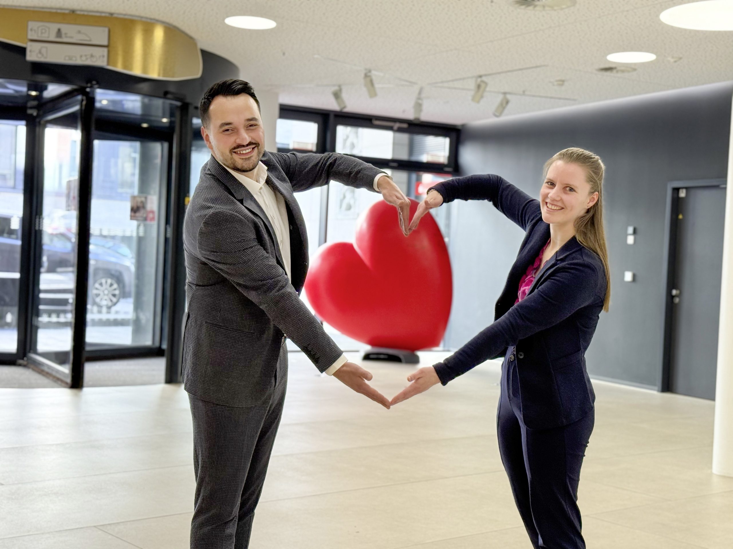 Carina Schmid und Okan Güldal stehen nebeneinander und formen ein Herz mit den Armen. In der Mitte des Herzes ist im Hintergrund ein rotes Holzherz zu erkennen. Der Bildausschnitt ist vom Eingangsbereich der Sparkasse.