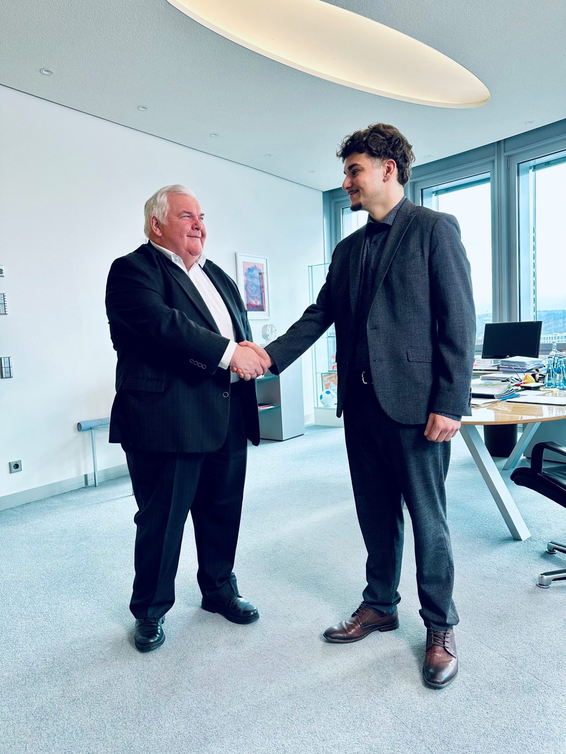 Hans Neuweiler schüttelt Kian Rüdenauer freundlich die Hand. Beide stehen und blicken einander an. Hintergrund ist in freundlicher Büroathmosphäre.