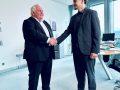 Hans Neuweiler schüttelt Kian Rüdenauer freundlich die Hand. Beide stehen und blicken einander an. Hintergrund ist in freundlicher Büroathmosphäre.