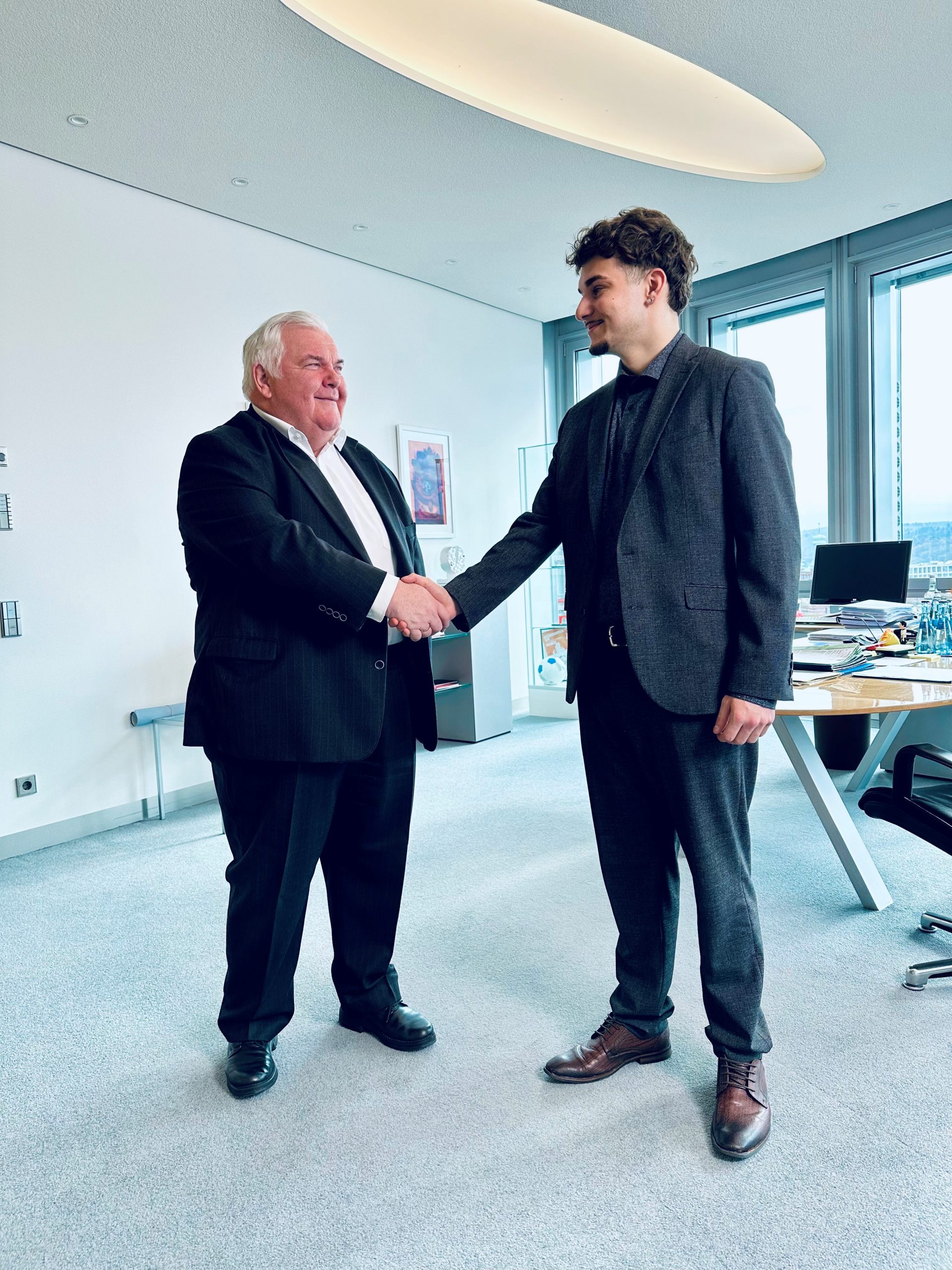 Hans Neuweiler schüttelt Kian Rüdenauer freundlich die Hand. Beide stehen und blicken einander an. Hintergrund ist in freundlicher Büroathmosphäre.