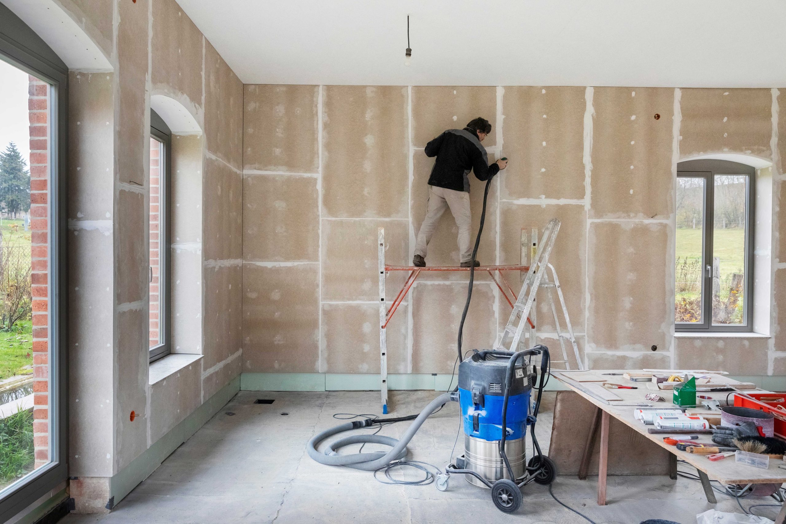Mann auf einer Bauleiter, der seine Wand renoviert. Im Vordergrund stehen ein Baustaubsauger und ein Malertisch mit reichlich Werkzeug. Der Raum befindet sich im Rohbau.