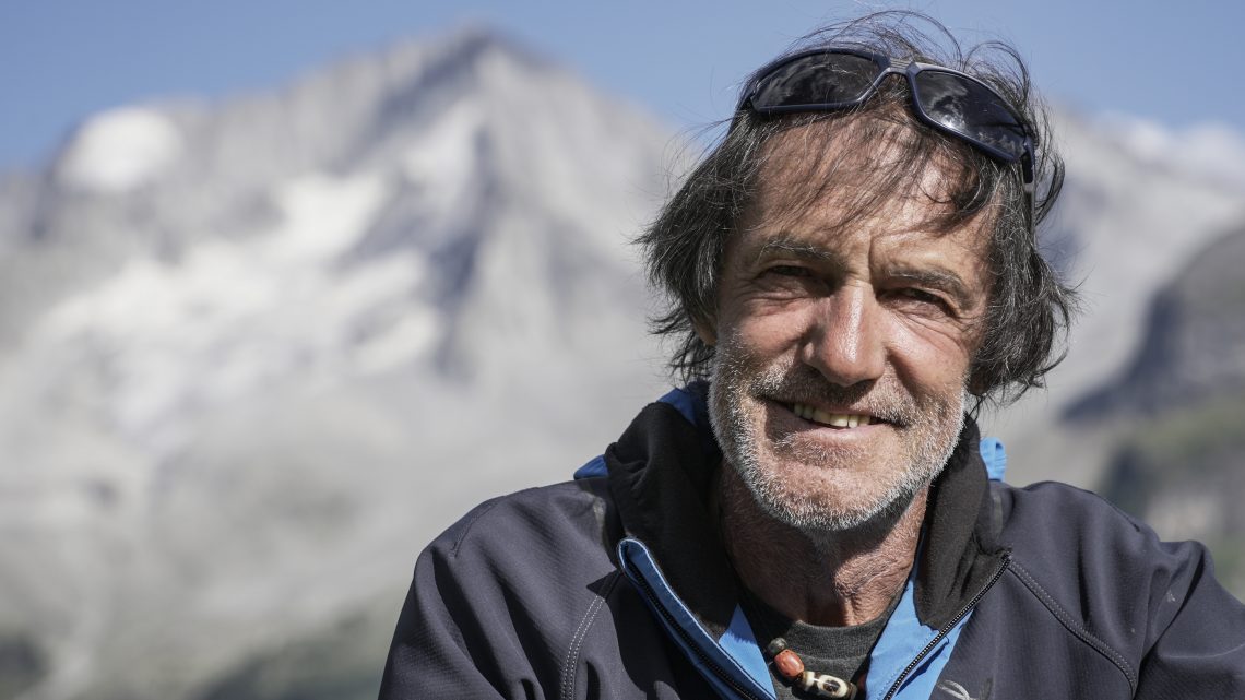 Profilfoto von Hans Kammerlander der freundlich in die Kamera lächelt. Im Hintergrund ein schneebedeckter Berg mit blauem Himmel