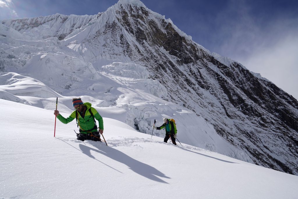 Zwei Männer stecken hüfttief in Schnee. Sie haben Walkingstöcke und erklimmen einen schneebedeckten Berg. Im Hintergrund ein schönes Bergpanorama. 