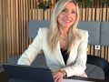 Frau Kristina Sauter sitzt am Laptop und lächelt freundlich in die Kamera. Vor ihr liegt ein Bauzeichnung. Im Hintergrund freundliche Umgebung mit Holzpanelen und grünen Zimmerpflanzen.