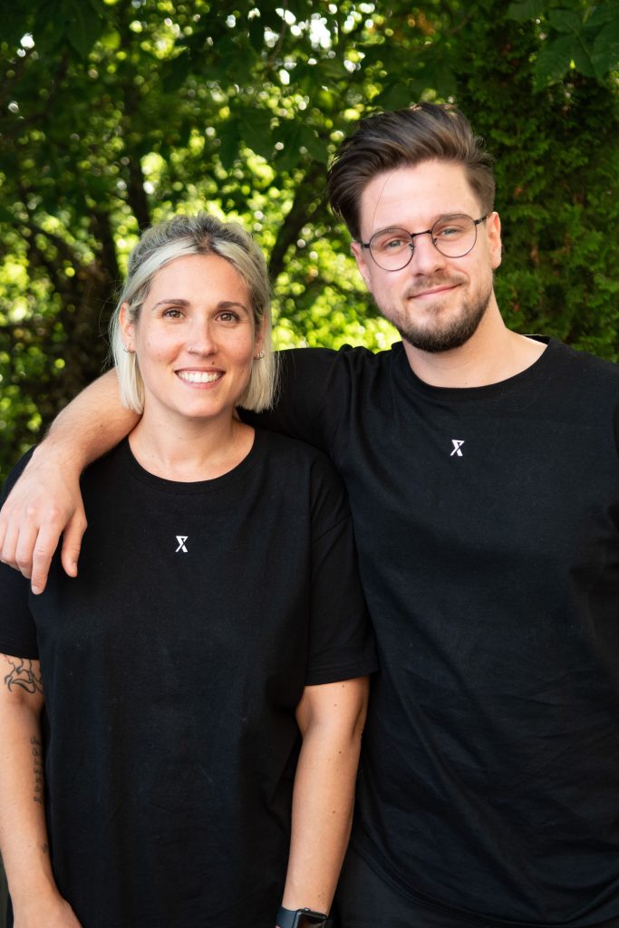Portraitbild von Lisa Dengler und Mark Hofmann. Beide tragen ein schwarzes PF Next-Shirt und lächeln freundlich in die Kamera. Im Hintergrund sind grüne Bäume zu erkenn. 
