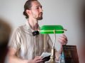 Tristan Krieg an einem Rednerpult, Eine grüne altmodische Schreibtischlampe steht bei ihm. Er liest sichtlich ergriffen aus seinem Buch vor.