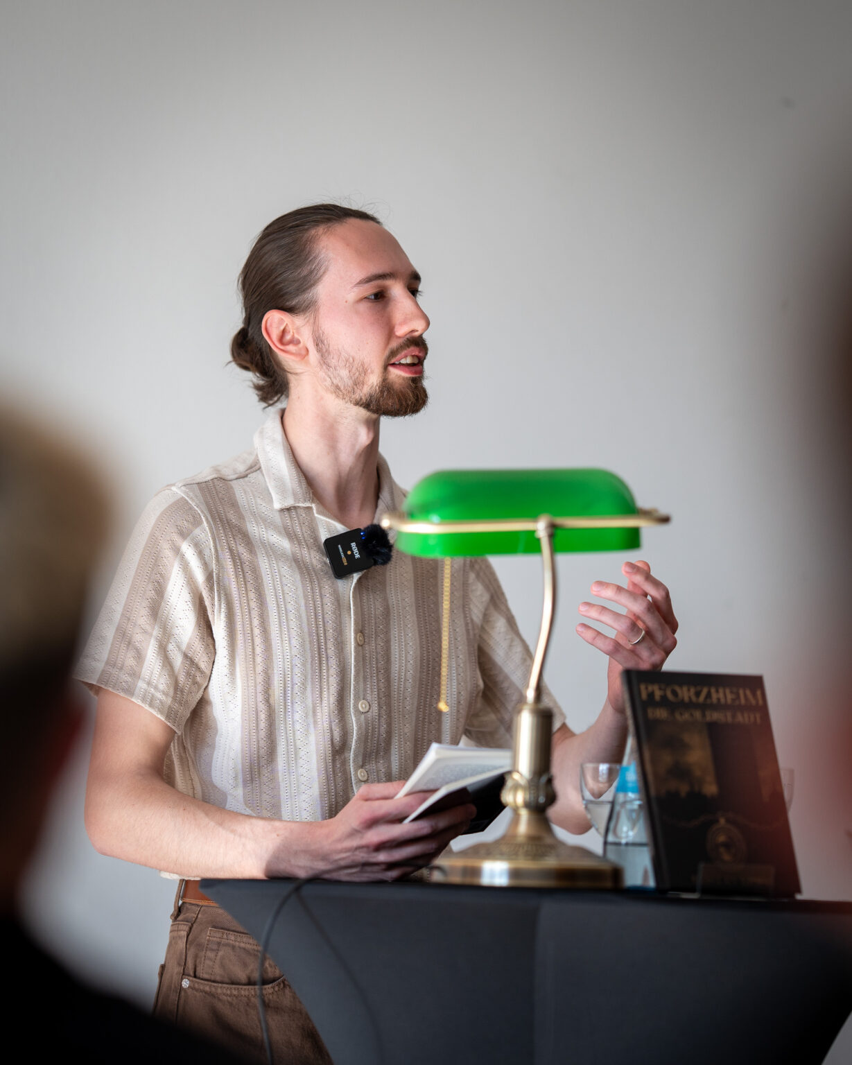 Tristan Krieg an einem Rednerpult, Eine grüne altmodische Schreibtischlampe steht bei ihm. Er liest sichtlich ergriffen aus seinem Buch vor.