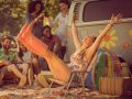 Mädchen sitzt vor VW Bus auf einem Campingstuhl. Sie streckt Arme und Beine freudig aus. Sie trägt quitsch bunte Gummistiefel. Im Hintergrund sind junge Leute, in entspannter Festival-Camping-Athmosphäre.