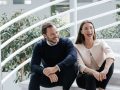 Geschwister Alicia und Nicolas Lindner sitzen nebeneinander auf einer Treppe. Das Bild ist hell und modern. Im Hintergrund sind grüne Pflanzen zu erkennen. Alicia Lindner lacht herzlich.