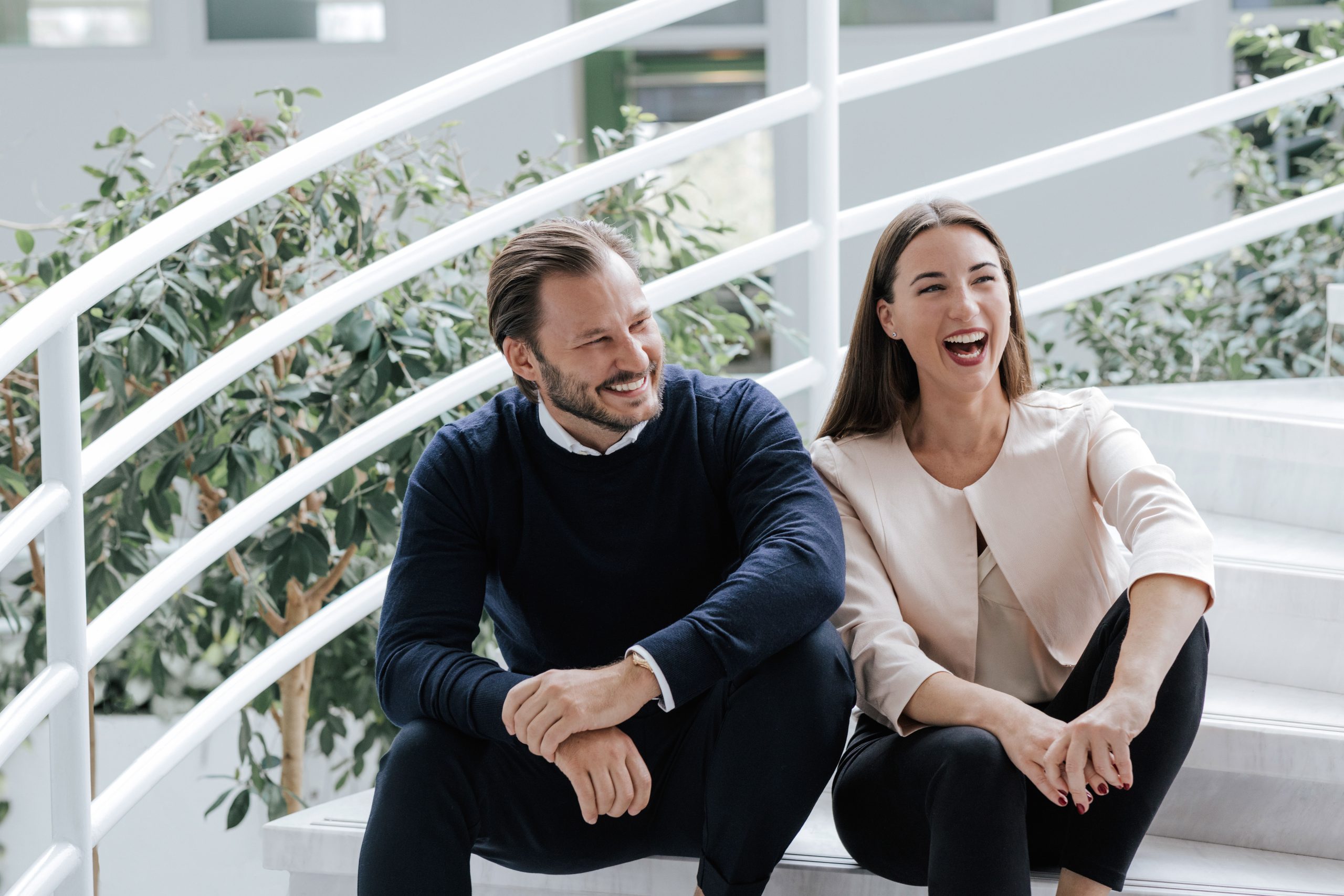 Geschwister Alicia und Nicolas Lindner sitzen nebeneinander auf einer Treppe. Das Bild ist hell und modern. Im Hintergrund sind grüne Pflanzen zu erkennen. Alicia Lindner lacht herzlich.